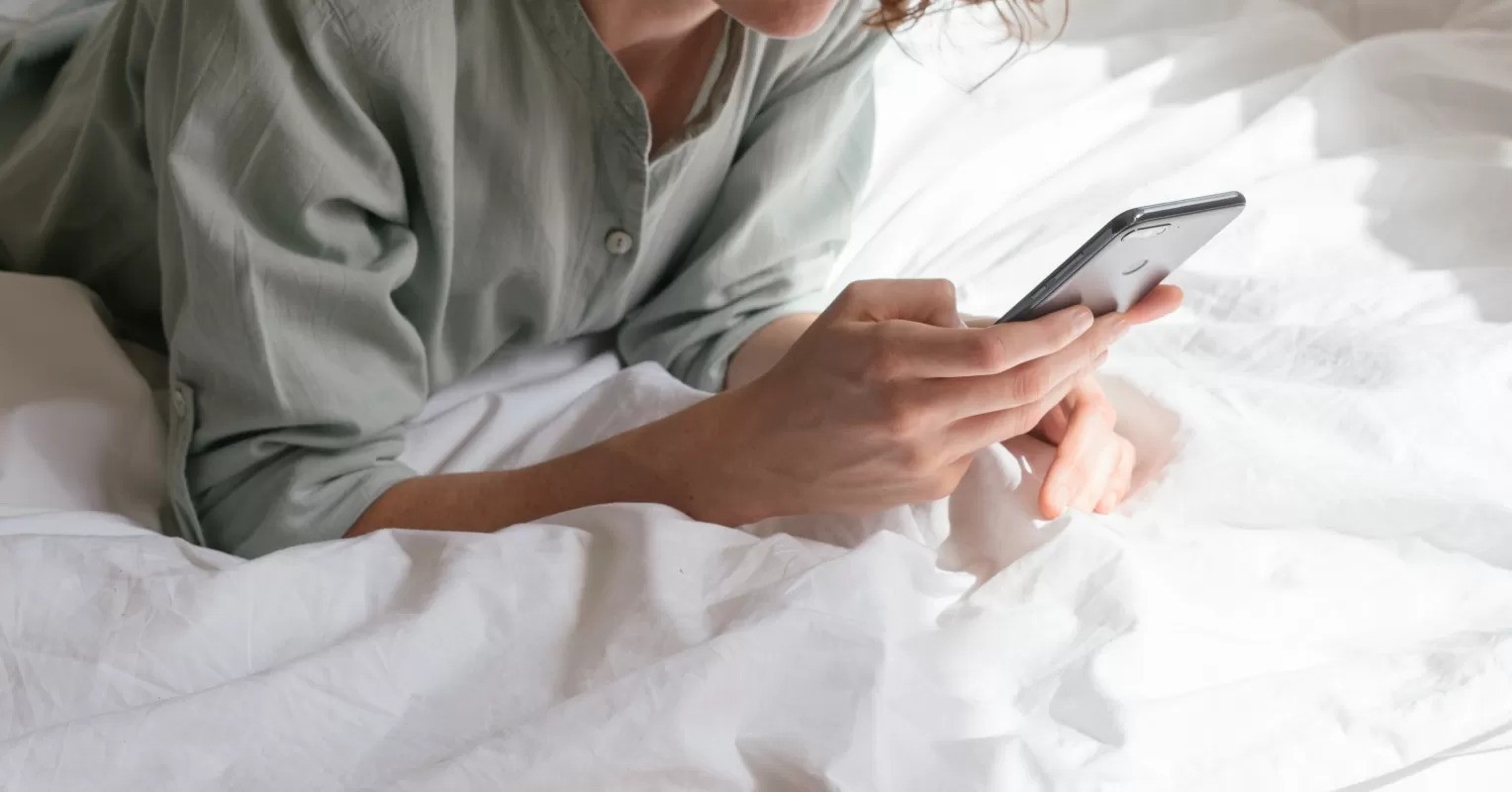 A lady typing on a smartphone while lying on a bed covered in white sheets, wearing a light green shirt