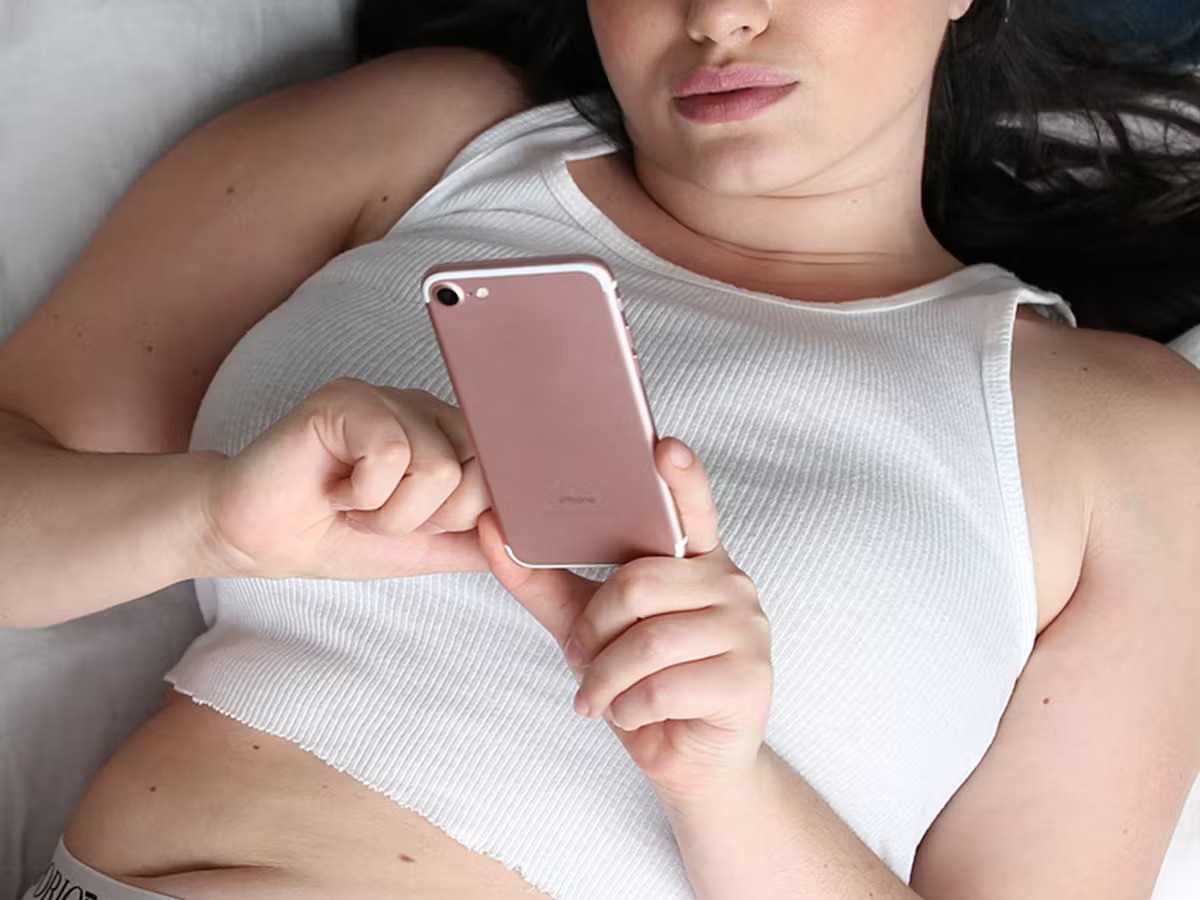 A person lies on their back, using a rose gold phone, wearing a white tank top